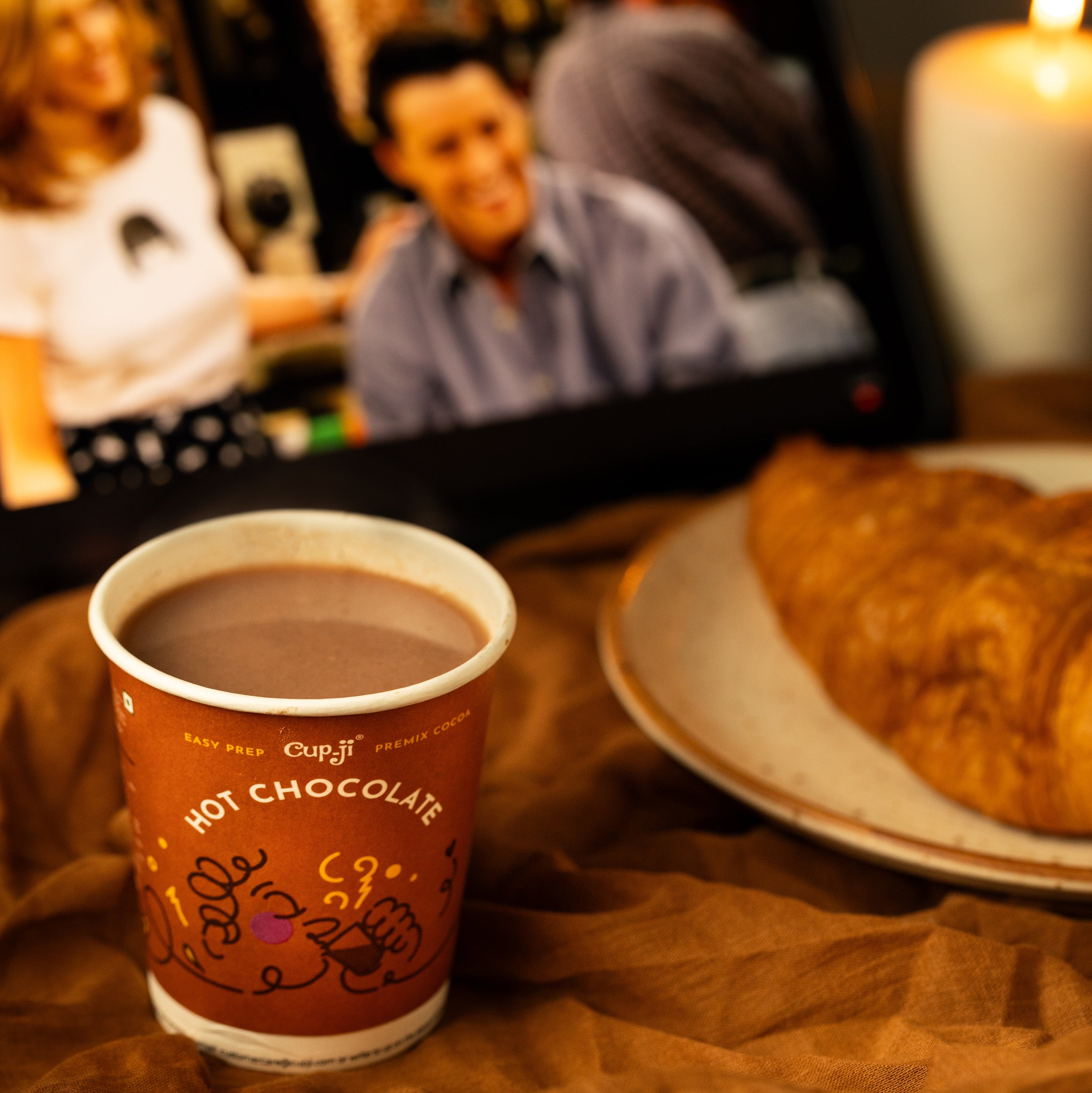 Hot chocolate cup with a croissant on a table, blurred background of people and a candle.