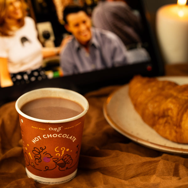 Hot chocolate cup with a croissant on a table, blurred background of people and a candle.